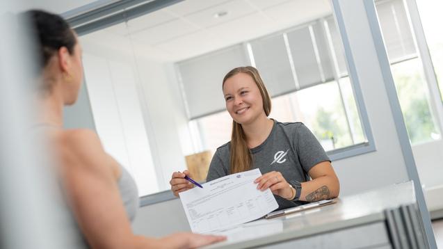 Zwei Frauen sitzen an einem Tisch; eine lächelt und zeigt auf ein Dokument, während die andere aufmerksam zuhört.