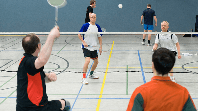 Vielzählige Personen im Badmintonspiel in einer Sporthalle, während ein Spieler den Ball schlägt.