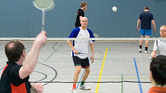 Vier Personen spielen Badminton in einer Sporthalle, w&auml;hrend ein Spieler den Federball schl&auml;gt.