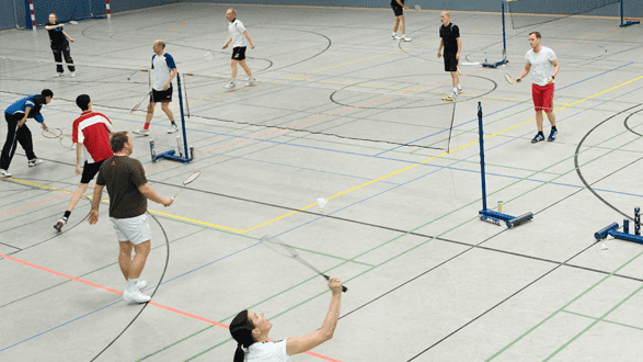 Mehrere Personen spielen Badminton in einer Sporthalle mit markierten Linien auf dem Boden.