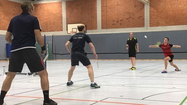 Fünf Personen spielen Badminton in einer Sporthalle. Ein Spieler schlägt den Ball über das Netz.