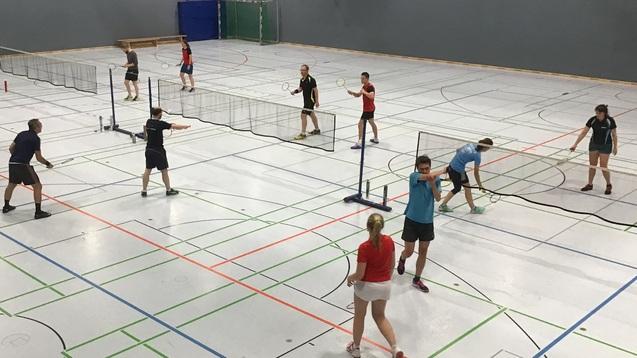 Mehrere Personen spielen Badminton in einer hellen Sporthalle mit Netz und markierten Spielfeldern.