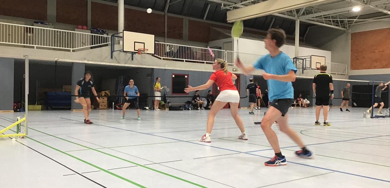Mehrere Personen spielen Badminton in einer Sporthalle, mit Netzen und Bällen sichtbar im Hintergrund.