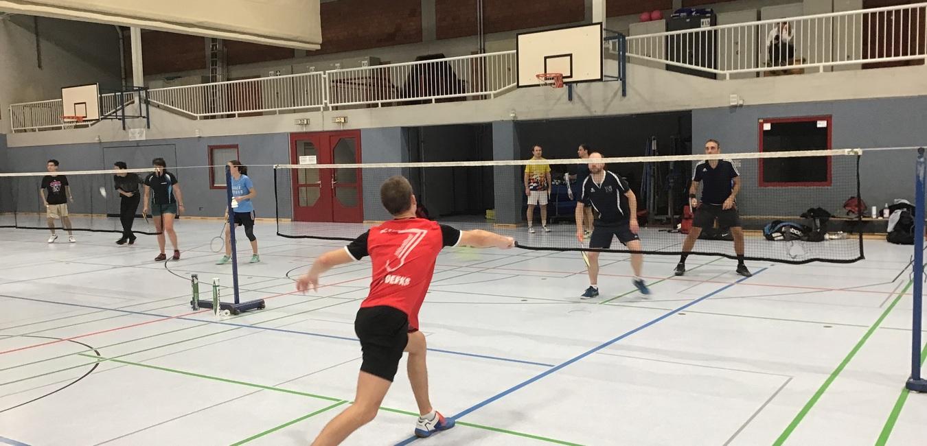 Badminton-Spiel in einer Sporthalle, Spieler schlägt den Federball, Zuschauer stehen an der Seite.