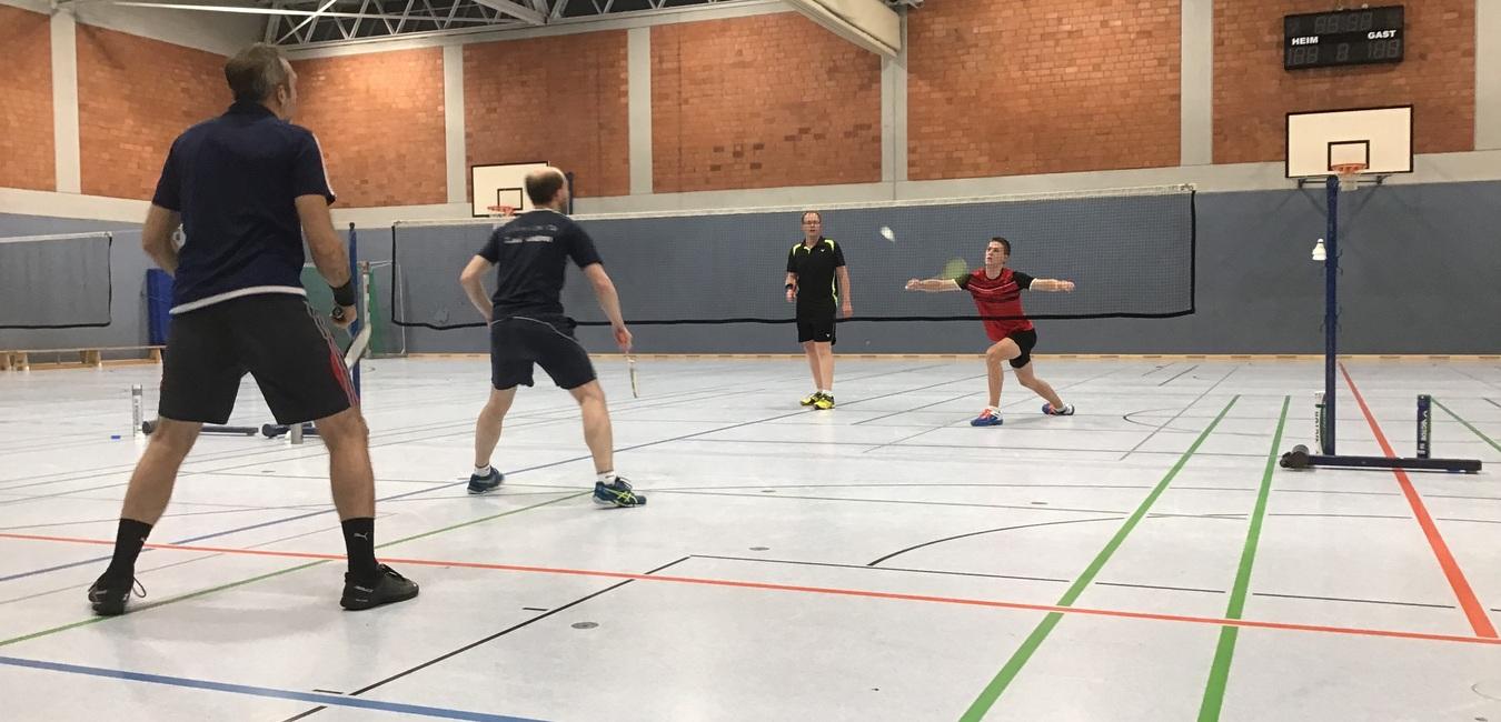 Fünf Personen spielen Badminton in einer Sporthalle. Ein Spieler schlägt den Ball über das Netz.
