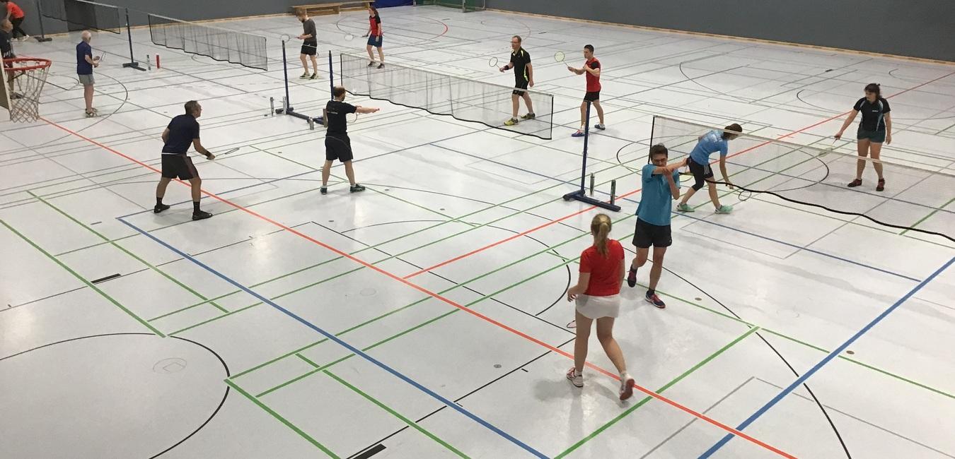 Mehrere Personen spielen Badminton in einer hellen Sporthalle mit Netz und markierten Spielfeldern.