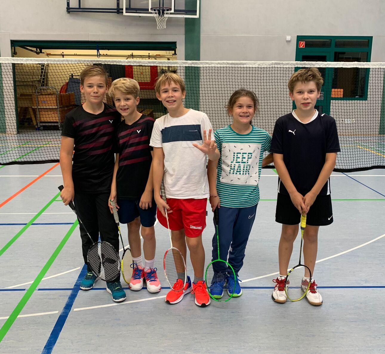 Fünf Kinder stehen in einer Sporthalle mit Badmintonschlägern in der Hand, Hintergrund mit Netz und Spielfeldmarkierungen.