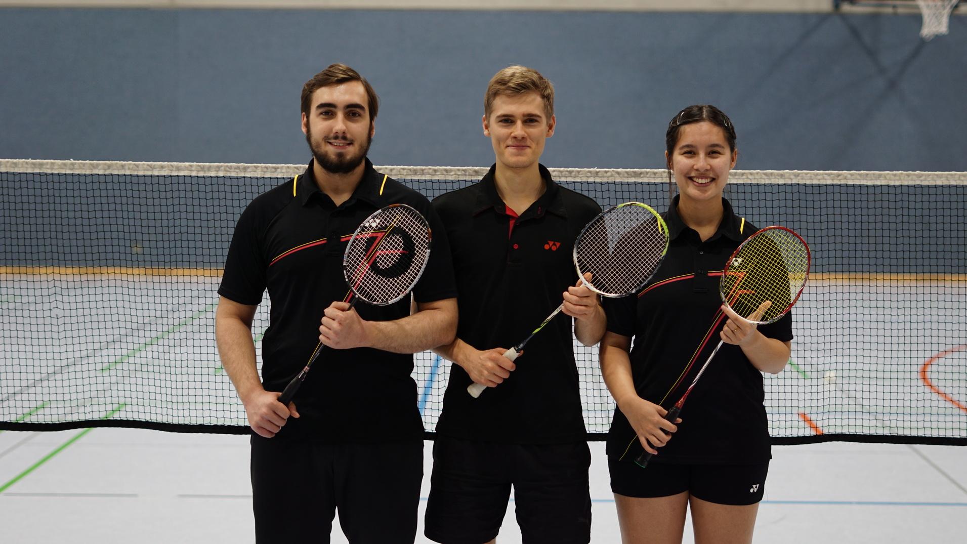 Drei sportliche Personen mit Badmintonschl&auml;gern stehen vor einem Badmintonnetz in einer Sporthalle.