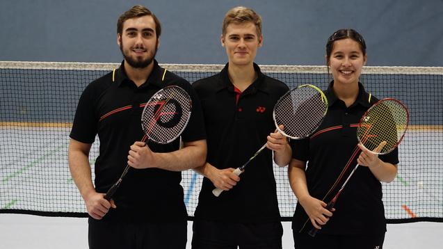 Drei sportliche Personen mit Badmintonschlägern stehen vor einem Badmintonnetz in einer Sporthalle.