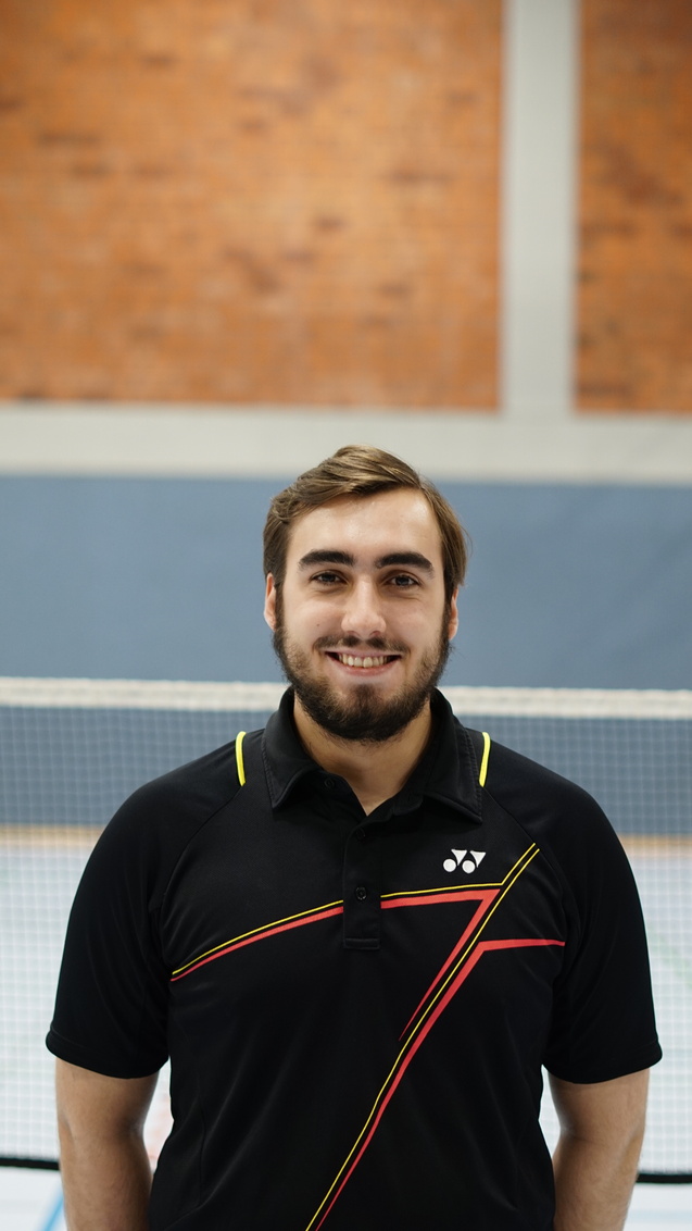 L&auml;chelnder junger Mann in schwarzem Sportshirt mit bunten Akzenten vor einem Tischtennisnetz in einer Halle.