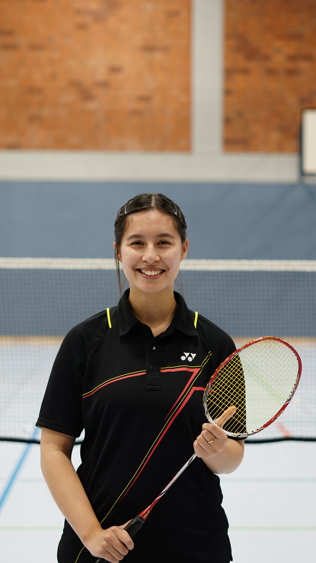 Junge Frau in Sportkleidung h&auml;lt einen Badmintonschl&auml;ger in einer Halle mit Badminton-Netz im Hintergrund.