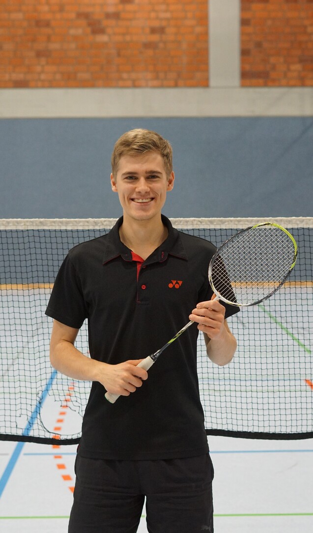 Junger Mann h&auml;lt einen Badmintonschl&auml;ger, steht vor einem Badmintonnetz in einer Sporthalle.