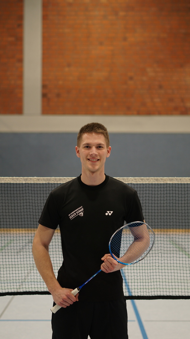 L&auml;chelnder junger Mann h&auml;lt einen Badmintonschl&auml;ger vor einem Badmintonfeld mit Netz und Ziegelwand im Hintergrund.
