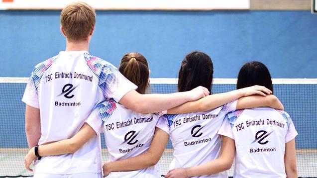 Vier Badmintonspieler stehen Arm in Arm, mit Rücken zur Kamera, in Teamtrikots des TSC Eintracht Dortmund.