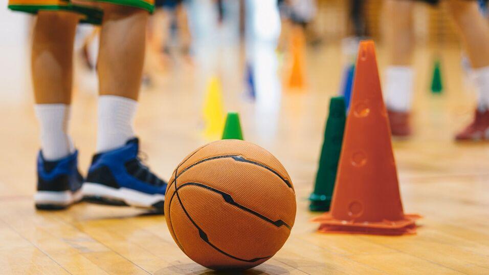 Nahaufnahme eines Basketballs auf einem Holzboden, umgeben von bunten Hütchen und Spielern im Hintergrund.