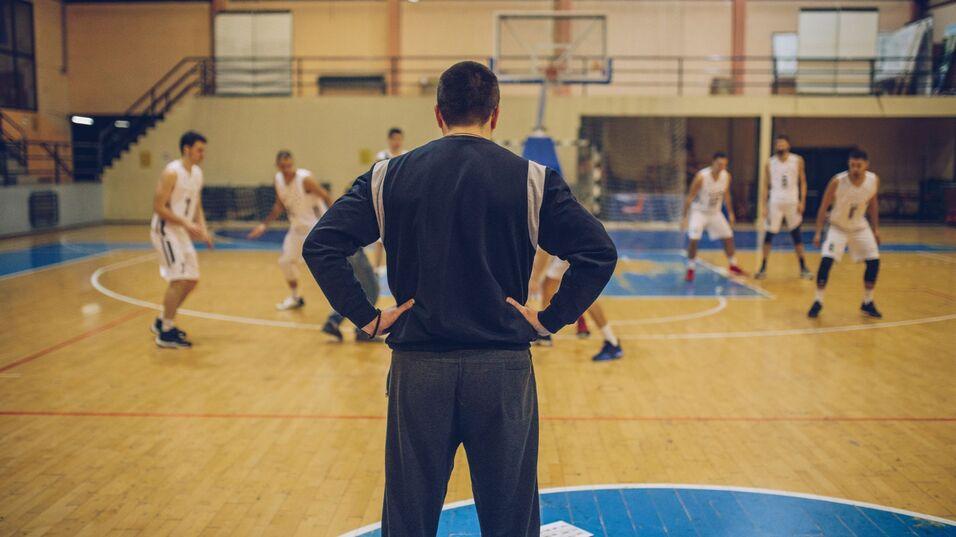 Eine Trainerperson beobachtet eine Basketballmannschaft beim Training auf dem Spielfeld.