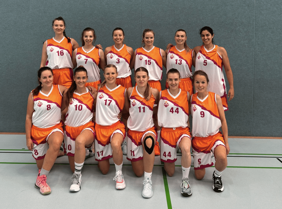 Gruppenfoto von zw&ouml;lf Basketballspielerinnen in orange-wei&szlig;en Trikots auf einem Sportplatz.