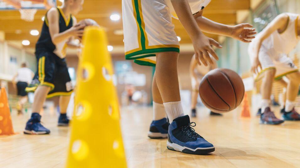 Junge dribbelt einen Basketball zwischen Trainingshütchen auf einem Holzsportboden.