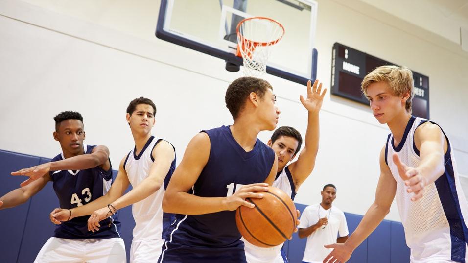 Fünf Basketballspieler in einer Sporthalle, einer mit Ball, während zwei Spieler versuchen, ihn zu verteidigen.