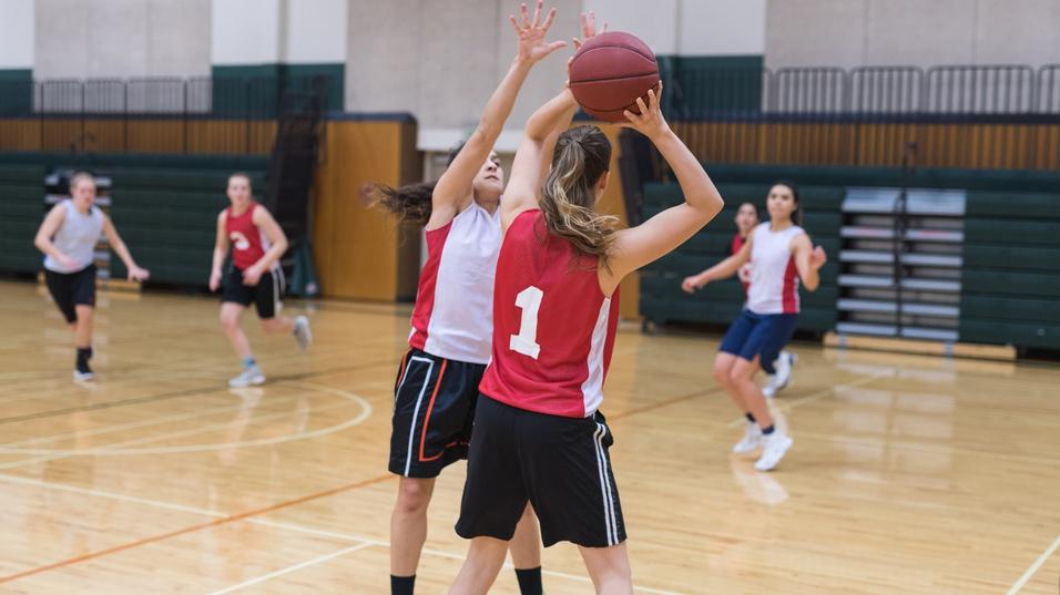 Zwei Basketballspielerinnen im Wettkampf, eine wirft den Ball, w&auml;hrend die andere sie blockt. Andere Spielerinnen im Hintergrund.