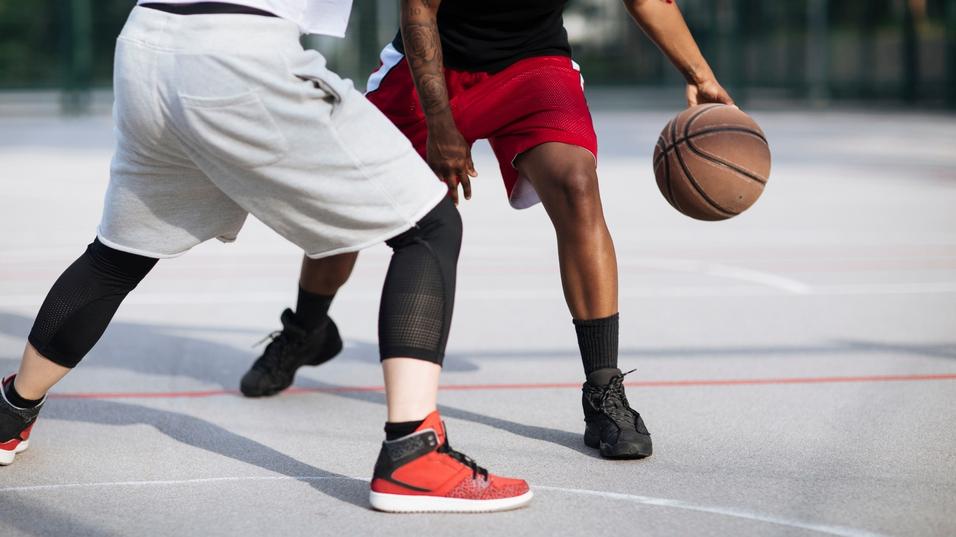 Zwei Spieler dribbeln einen Basketball auf einem Sportplatz, der Asphalt ist markiert und gut beleuchtet.