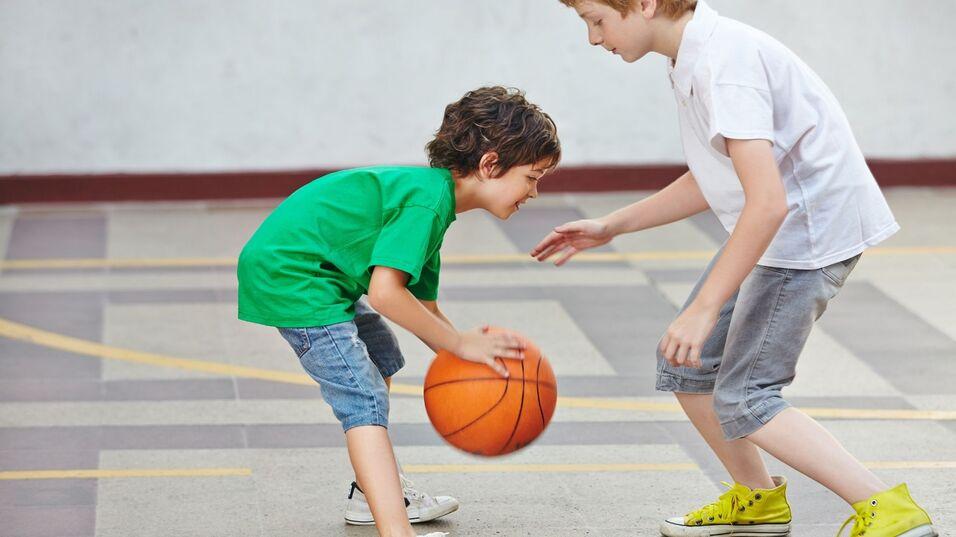Zwei Kinder spielen Basketball in einer Sporthalle, eines dribbelt den Ball, während das andere zuschaut.