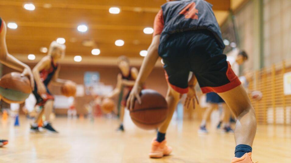 Mehrere Jugendliche dribbeln Basketballs auf einem Parkettboden in einer Halle während des Trainings.