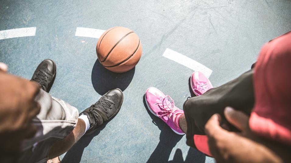 Nahaufnahme von zwei Paar F&uuml;&szlig;en auf einem Basketballplatz mit einem Basketball in der Mitte.