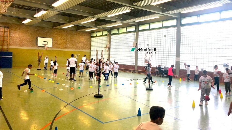 Schüler in weißen T-Shirts absolvieren ein Sportevent in einer Turnhalle mit verschiedenen Übungsstationen.