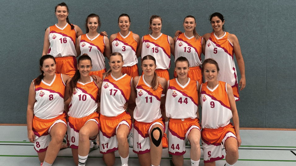 Gruppenfoto von zwölf Basketballspielerinnen in orange-weißen Trikots auf einem Sportplatz.