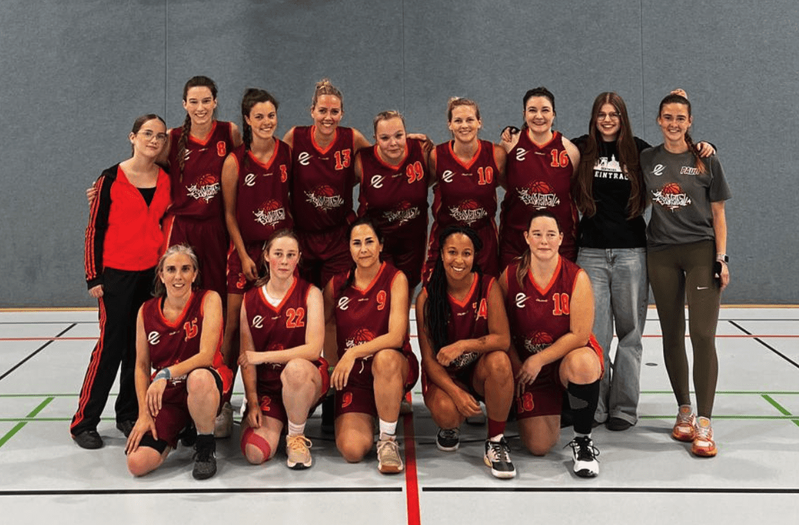 Gruppenfoto eines Frauen-Basketballteams in roten Trikots auf einem Sportplatz, aufgestellt in zwei Reihen.