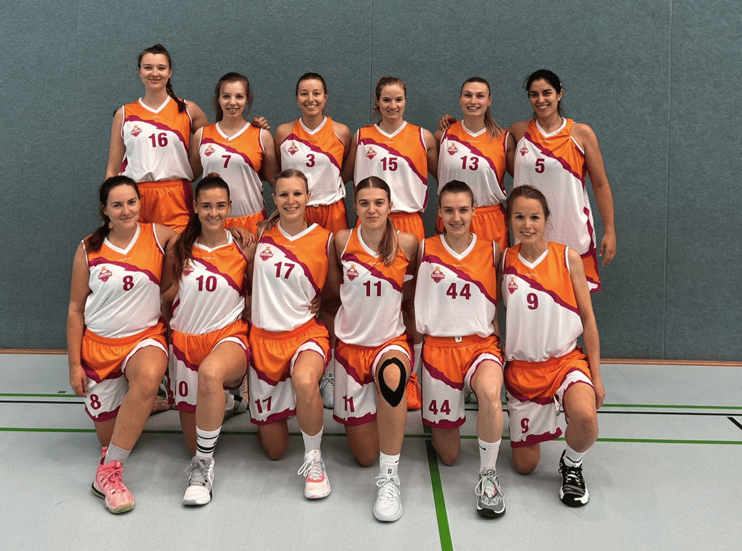 Gruppenfoto von zw&ouml;lf Basketballspielerinnen in orange-wei&szlig;en Trikots auf einem Sportplatz.