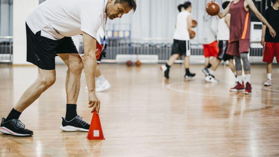 Trainer platziert ein rotes Kegelsignal auf dem Holzboden eines Basketballfeldes, während Spieler im Hintergrund trainieren.