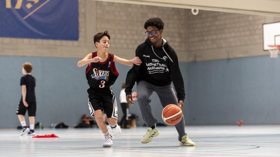 Ein Trainer dribbelt mit einem Basketball, w&auml;hrend ein Kind in Basketballkleidung ihm nachl&auml;uft.