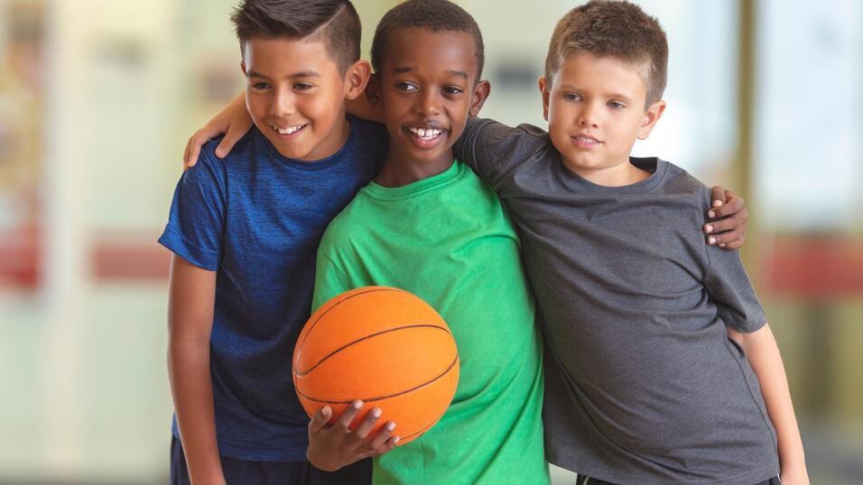 Drei Jungen stehen eng zusammen und halten einen Basketball, lächelnd in sportlicher Kleidung.