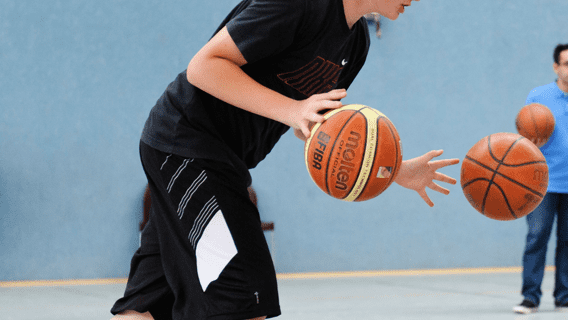 Junger Spieler dribbelt zwei Basketballs in einer Sporthalle, w&auml;hrend andere Spieler im Hintergrund warten.