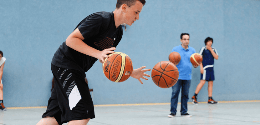 Junger Spieler dribbelt zwei Basketballs in einer Sporthalle, während andere Spieler im Hintergrund warten.