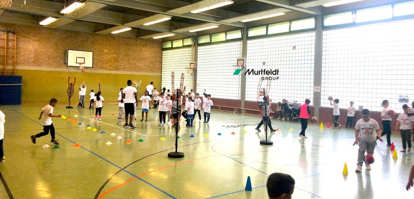 Schüler in weißen T-Shirts absolvieren ein Sportevent in einer Turnhalle mit verschiedenen Übungsstationen.