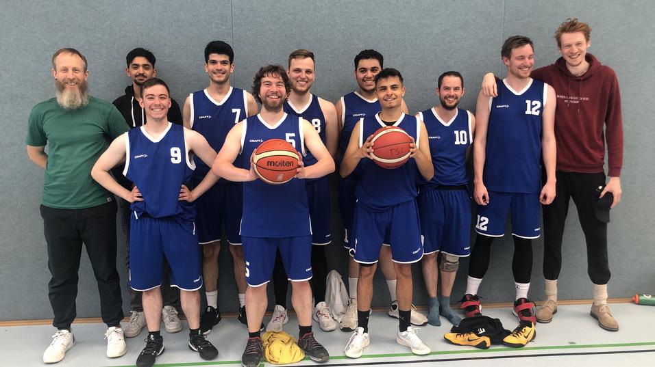 Eine Basketballmannschaft mit zehn Spielern in blauen Trikots posiert l&auml;chelnd auf einem Sportfeld.