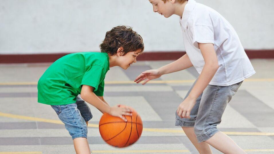Zwei Kinder spielen Basketball in einer Sporthalle, eines dribbelt den Ball, w&auml;hrend das andere zuschaut.