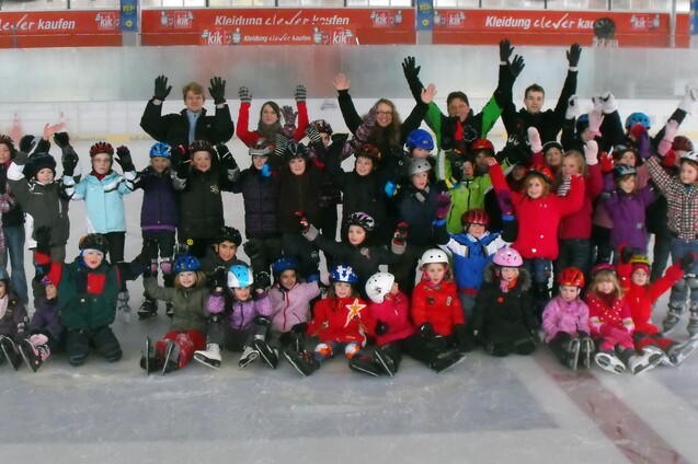 Gro&szlig;e Gruppe von Kindern in Winterkleidung mit Schlittschuhen auf einer Eisbahn, alle heben die H&auml;nde in die Luft.
