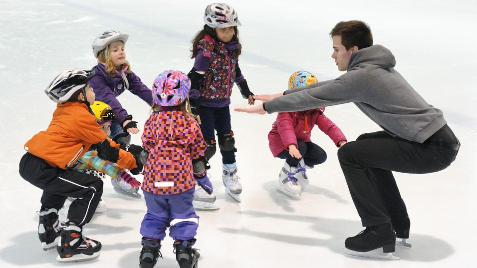 Truppe von Kindern in Eishockeyausr&uuml;stung um einen Trainer, der ihnen das Eislaufen auf einer Eisfl&auml;che zeigt.