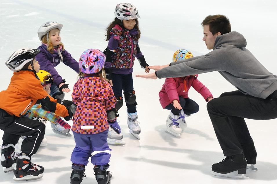 Truppe von Kindern in Eishockeyausrüstung um einen Trainer, der ihnen das Eislaufen auf einer Eisfläche zeigt.