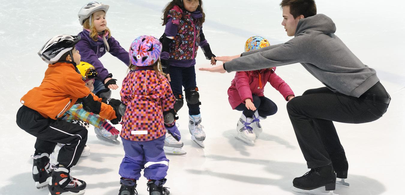 Truppe von Kindern in Eishockeyausr&uuml;stung um einen Trainer, der ihnen das Eislaufen auf einer Eisfl&auml;che zeigt.