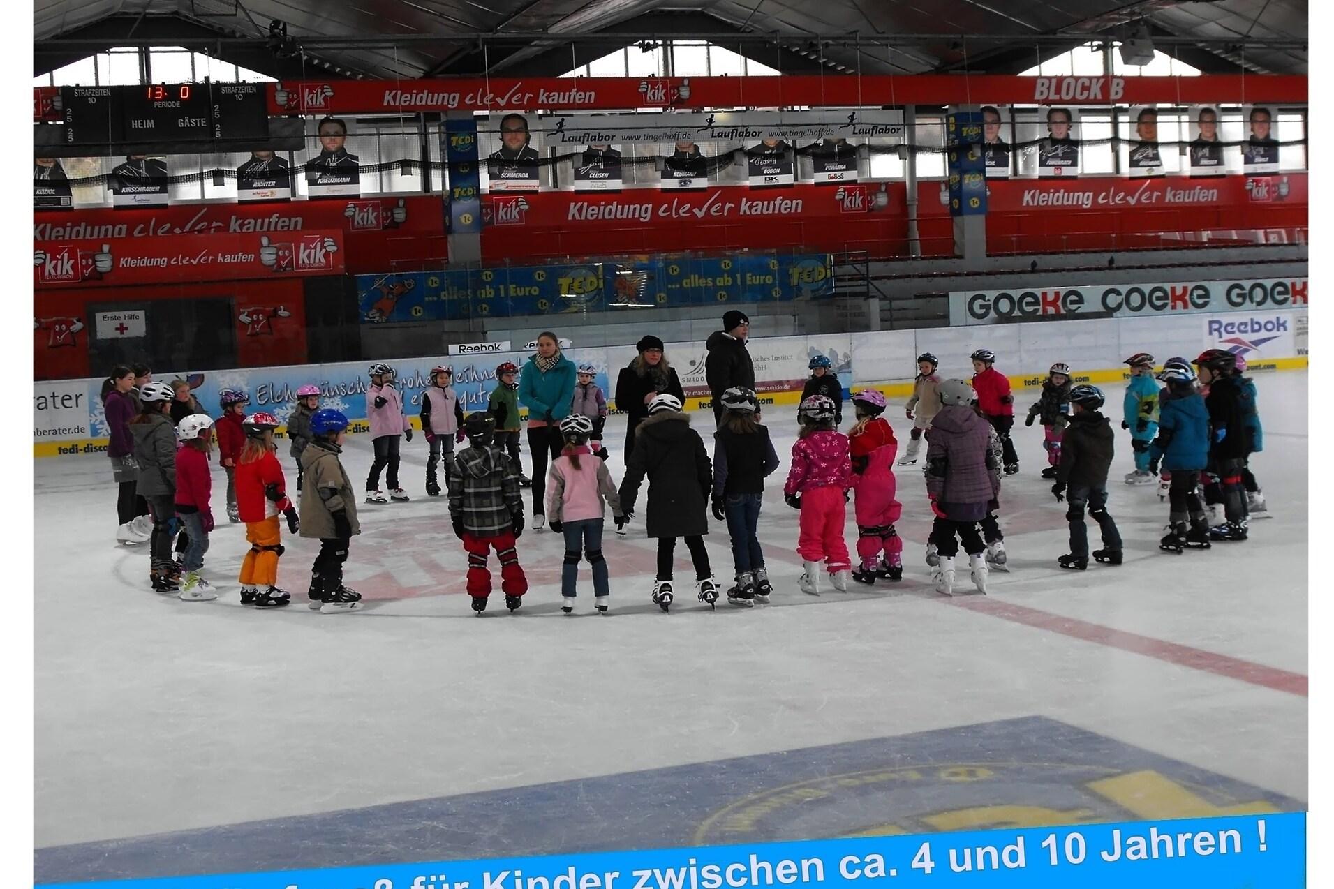 Gruppe von Kindern in Eislaufschuhen auf einer Eisbahn, umgeben von Betreuern, während eines Eislaufkurses.