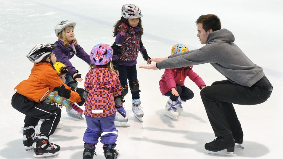 Truppe von Kindern in Eishockeyausr&uuml;stung um einen Trainer, der ihnen das Eislaufen auf einer Eisfl&auml;che zeigt.
