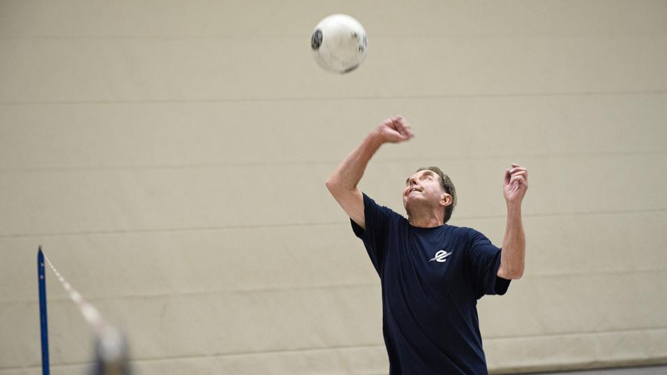 Mann in Sportkleidung hebt einen Ball zur Annahme in einer Sporthalle mit Netz im Hintergrund.