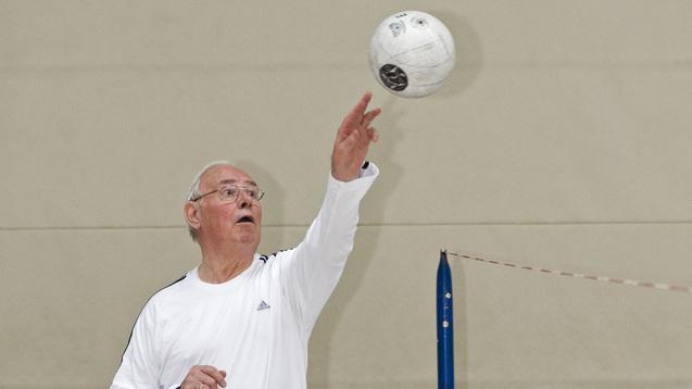 Ein älterer Mann spielt Volleyball und wirft einen weißen Ball in einer Sporthalle.