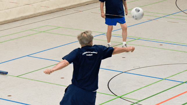 Drei M&auml;nner spielen Volleyball in einer Sporthalle; ein Mann schl&auml;gt einen Ball &uuml;ber das Netz.