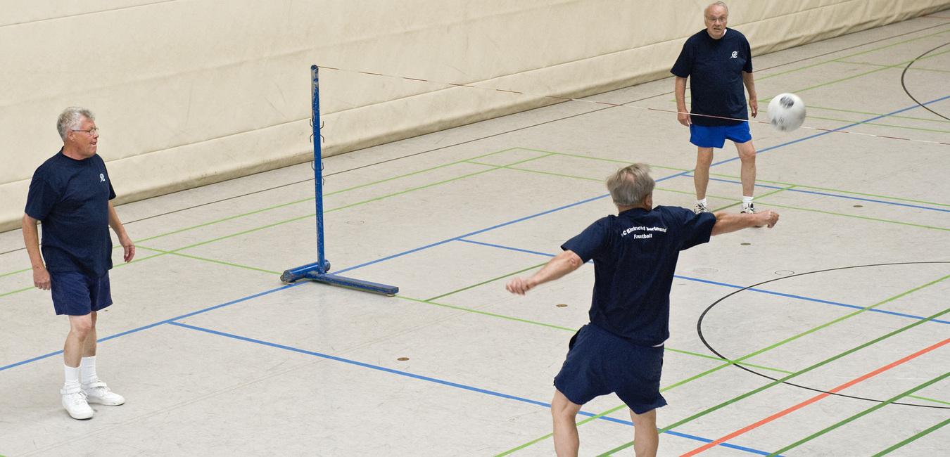 Drei Männer in Sportkleidung spielen Volleyball in einer Sporthalle mit Netzkonstruktion und markierten Spielfeldern.